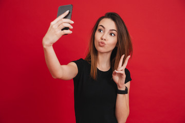 Lovely woman in black t-shirt having fun and taking selfie with gesturing peace sign on her smartphone, isolated over red background