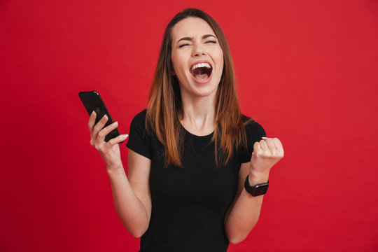 Portrait of ecstatic woman 20s wearing black t-shirt screaming and clenching fist like rejoicing victory or triumph, isolated over red background
