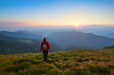 Redheaded girl athlete with a backpack and sticks stands on the green hillocks and looks at high mountain landscapes and fascinating sunset. Location place Carpathian national park, Ukraine, Europe.