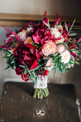 Bridal bouquet in wine red color with lace ribbon on wooden chair, wedding rings near