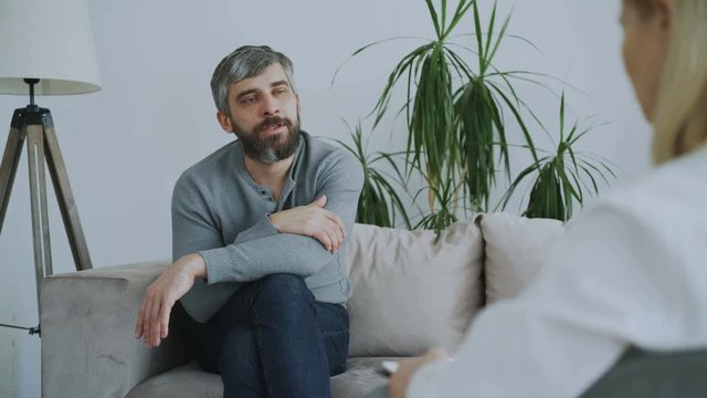 Adult bearded man sitting on couch talking to female psychotherapist in office indoors