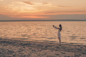 Young girl stands on the beach and takes a picture of the sunset on her smartphone