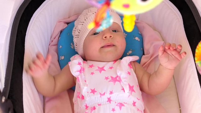Active three months baby girl in crochet summer hat and pink onesie lying in pram with toys hanging above