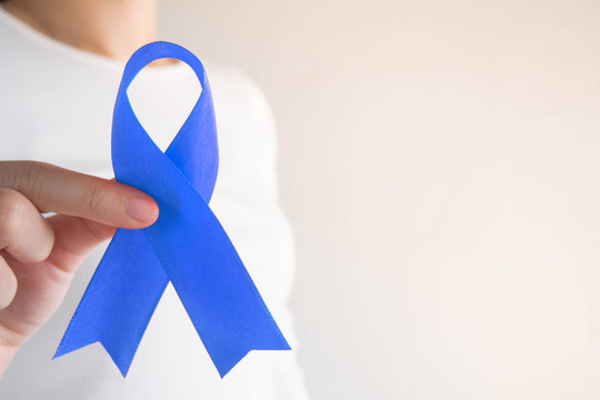 Dark blue ribbon awareness on female hands in white t-shirt background w/ copy space. Symbol for support people living w/ colon or colorectal cancer. Health care and medical concept.