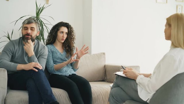 Young couple visiting professional psychologist office. Curly woman angrily arguing and talking about her boyfriend to marriage counselor