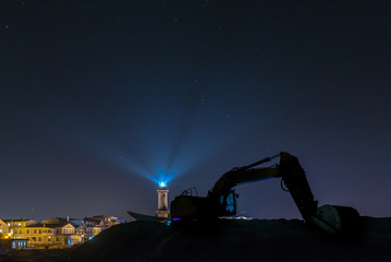 Silhouette of an excavator in front of the landmark Leuchtturm Warnem&uuml;nde at night. Concept: maritime or vacation and travel