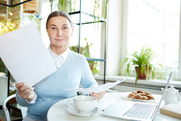 Contemporary mobile businesswoman with earphones holding financial documents while working by table...