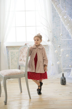Adorable cute girl in red dress and pink coat is stayng near the chair in a fairy white room
