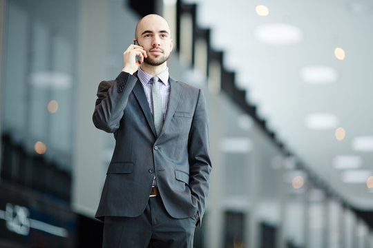 Young elegant businessman in suit speaking by smartphone in airport during travel