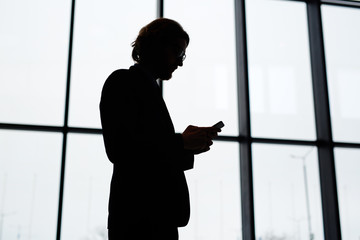Silhouette of contemporary businessman with smartphone messaging by airport window