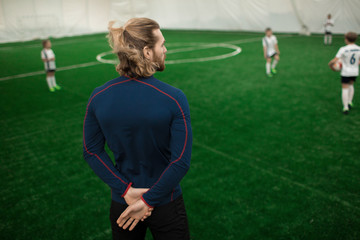 Rear view of football trainer in activewear standing on field and watching for players