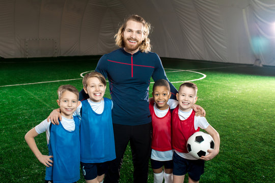Successful football trainer and four happy boys in uniform standing on green soccer field