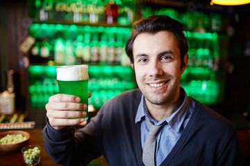 Young man with toothy smile holding glass of foaming beer while spending time in pub