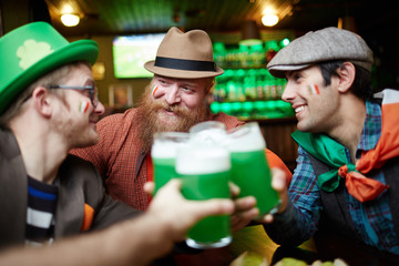 Happy young companions sitting in pub and toasting with green beer in glasses