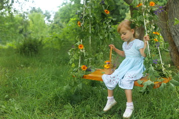 Happy little blonde caucasian child girl in light blue dress and roses in hair swing on swings in a meadow full of plants red roses yellow chamomiles and dandelions in spring