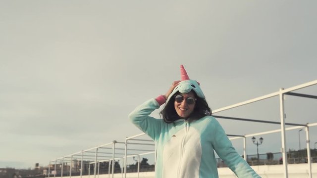 Joyful young woman is jumping on a pebble beach. She is dancing in kigurumi costume of unicorn and looking in camera