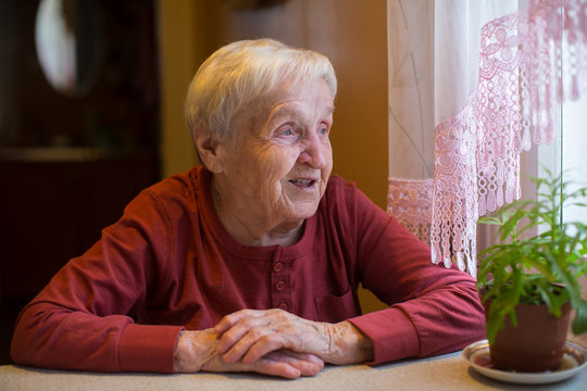 An elderly woman looks out the window sitting at the table.