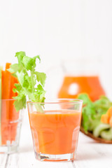 Carrot juice in beautiful glasses, cut carrot and green celery on wood bark bowl on white wooden background. Fresh vegetable drink. Close up photography. Selective focus. Horizontal banner
