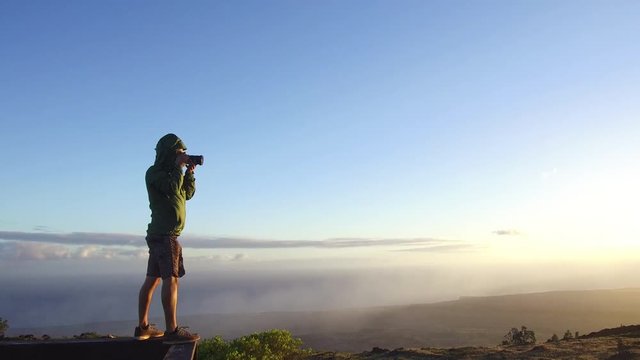 A young, attractive mixed race male photographer takes photos from a lookout at sunset, using a DSLR camera. His dark green jacket blows in the wind as he snaps photos of the setting sun. 