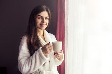 Beautiful girl wears white bathrobe holding cup of coffee near window