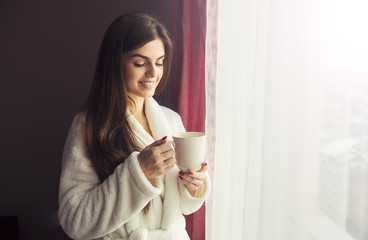 Beautiful girl wears white bathrobe holding cup of coffee near window