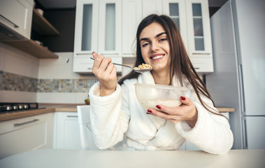 Beautiful girl wears white bathrobe eating cornflakes with milk in the kitchen in the morning