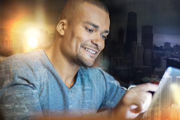 Feeling glad. Cheerful emotional handsome man sitting at the table and smiling while looking at the screen of a modern tablet