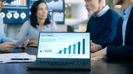 In the Meeting Room Laptop Standing on the Conference Desk Shows Company's Annual Growth Report with Interactive Charts. In the Background Business People Have Important Discussion.