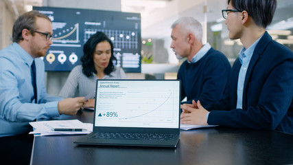 Obraz premium In the Meeting Room Laptop Standing on the Conference Desk Shows Company's Annual Growth Report with Interactive Charts. In the Background Business People Have Important Discussion.
