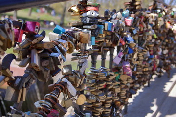 Ogromna ilość różnokolorowych kłódek symboli miłości zawieszonych na balustradzie mostu, część nieostra, w tle nieostry fragment parku, słonecznie