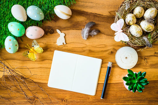 Leerer Notizblock mit Stift auf Holztisch im Hintergrund und bunte verzierte Ostereier, Federn, Schmetterlinge von oben zu Ostern