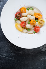 White plate with italian gnocchi, fresh tomatoes and green peas, above view on a grey concrete background with space, vertical shot
