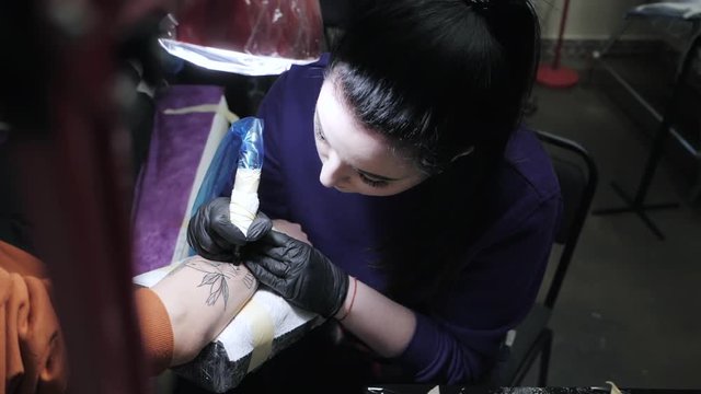 Young brunette female tattooer making tattoo on the man's arm of her client with gun. Slow motion. Dolly shot.