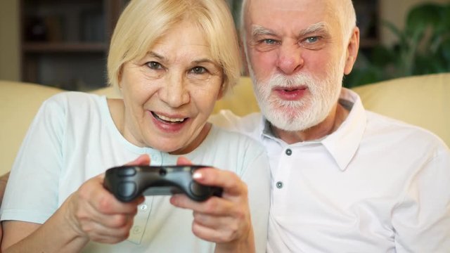 Cheerful senior couple gamers playing video games at home. Addicted seniorplayers with remote controller of game console. Cybersport for active modern elderly people