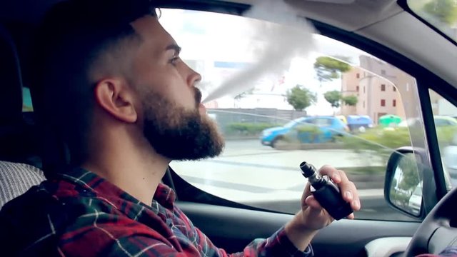 Young man with full beard smoking electronic cigarette while driving in the city. Hipster on checkers shirt with vaper pen in the car. Addiction, bad habit, quit smoking alternative concept