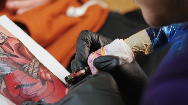 Professional master is painting tattoo with red ink, fills drawing with color. Works in black latex gloves with handmade rotor gun machine in studio. Slow motion.