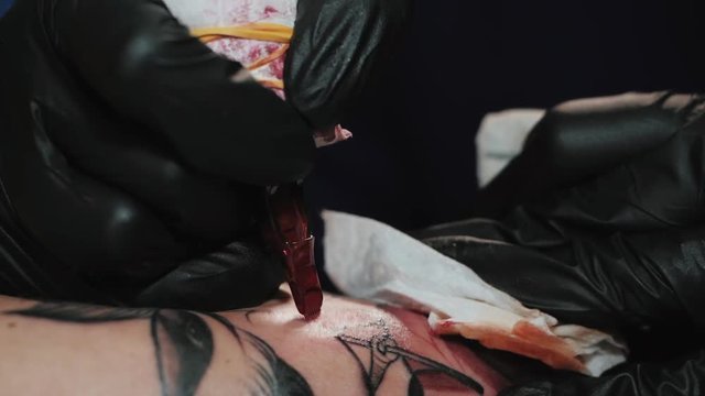 Professional master is painting tattoo with red ink, fills drawing with color. Works in black latex gloves with handmade rotor gun machine in studio. Slow motion.