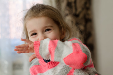a little girl with brown glistening eyes in a gray-white-pink sweater laughs and embarrasses covers her face with her hand