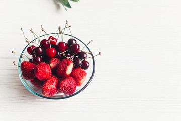 hello summer. fresh cherries and strawberries in stylish glass bowl  on white rustic wooden background. ripe juicy red  berries on table, harvest concept. space for text