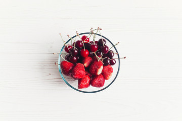 fresh cherries and strawberries in stylish glass bowl on white rustic wooden background. ripe juicy red mixed berries on table, harvest concept. space for text.  summer flat lay