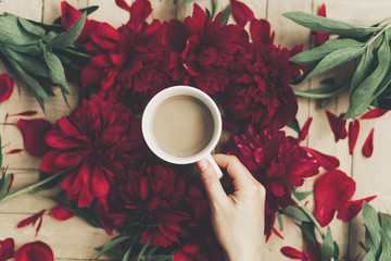 hand holding stylish coffee and beautiful red peonies on rustic wooden background flat lay. space for text. modern floral instagram blogging image. happy womens day or mothers
