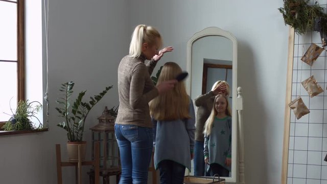 Positive single mother brushing amazing long blonde hair of cute little daughter while standing in front of the mirror. Loving mom combing her daughter's hair and chatting in domestic room.