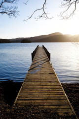 Fototapeta na wymiar very long wooden jetty, jutting out into a calm blue wooden lake with mountains in the background