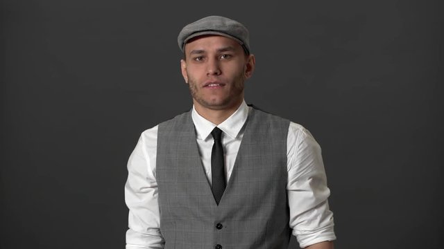 Portrait of innocent mature guy in vest and peaky kepi throwing up hands like he doesn't know or can't help over gray background. Concept of emotions