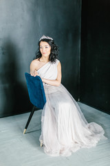 Young beautiful bride with a beautiful diadem in the hair poses sitting on the chair on dark background.