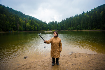 Happy elderly woman enjoying beautiful wiev of big mountain lake. Senior tourist woman near the forest lake taking selfie on action camera, harmony concept