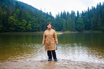 Happy elderly woman enjoying beautiful wiev of big mountain lake. Senior tourist woman near the forest lake taking selfie on action camera, harmony concept