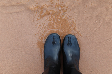 Black rubber boots on the sand top view. Feet in rubber boots in water