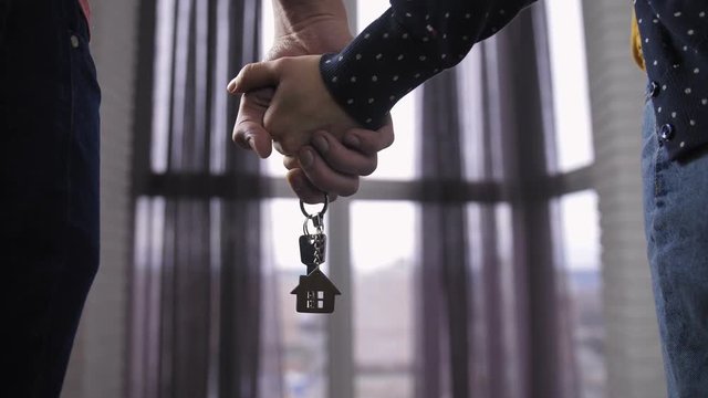 Close-up of couple standing in front of the window, holding hands with keys to their new home. Happy family purchased new apartment. Keys with wooden house shaped key ring.