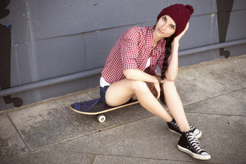 Brunette teenage girl in hipster outfit (jeans shorts, keds, plaid shirt, hat) with a skateboard at the park outdoors